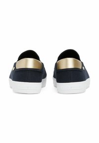 Scarpe slip-on in tela blu navy con un accento metallico oro sul tallone e suole in gomma bianca. La vista posteriore mostra il marchio sulla suola.