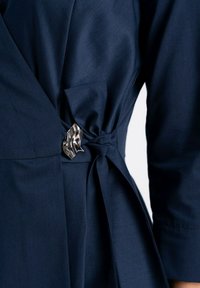Granatowa sukienka o kroju kopertowym, wykonana z gładkiego materiału, z wiązanym w pasie elementem i kontrastującą metaliczną broszką. Charakteryzuje się swobodną sylwetką.
