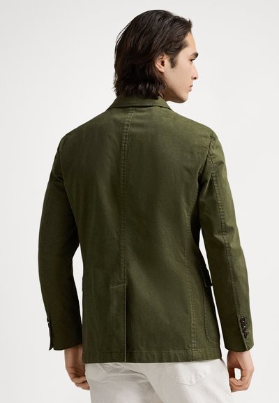 Χακί βαμβακερό μπουφάν με εφαρμοστή γραμμή, γιακάδες με εγκοπές και δύο πίσω σκισίματα· διαθέτει κουμπωτά μανίκια και πλαϊνές τσέπες.