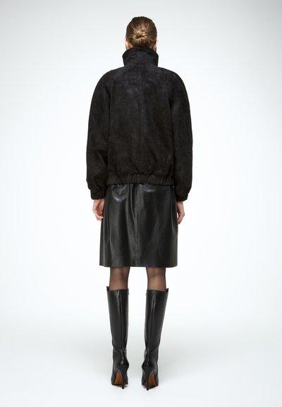 Femme avec un chignon portant une veste en daim noire, une jupe en cuir noire longue jusqu'aux genoux, des collants transparents et des bottes noires à talons montant jusqu'aux genoux, debout de dos.