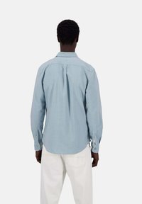 Camicia azzurro chiaro con maniche lunghe, piega sul retro, orlo arrotondato e polsini con bottoni, indossata con pantaloni bianchi.