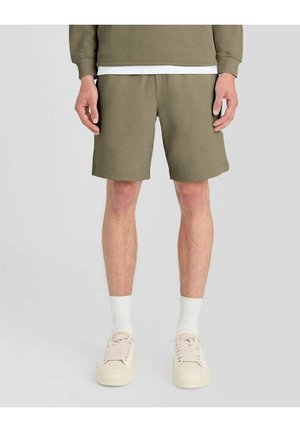 Man draagt olijfgroene shorts, witte sokken en off-white sneakers, staand tegen een effen lichte achtergrond.
