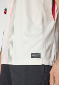 Maglietta sportiva bianca con strisce verticali verdi e rosse, tessuto strutturato, etichetta nera con il marchio "ENGINEERED" sul lato inferiore.