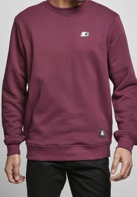 Hombre vistiendo un suéter de cuello redondo liso de color púrpura oscuro con un pequeño logo de estrella blanca en el pecho y una etiqueta negra cerca del dobladillo, combinado con pantalones negros.