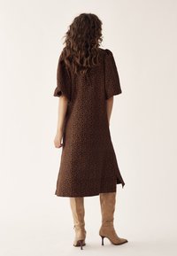 Femeie cu păr lung și creț, purtând o rochie midi maro cu motive florale și cizme maro deschis cu toc înalt, stând cu fața spre spate pe un fundal simplu.