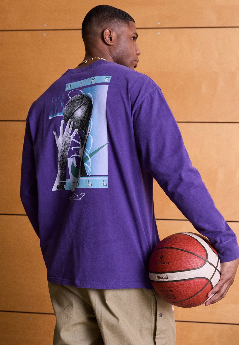 Maglietta a maniche lunghe viola con una stampa grafica di un pallone da basket e una mano sul retro. Il modello tiene in mano un pallone da basket, indossando pantaloni khaki.