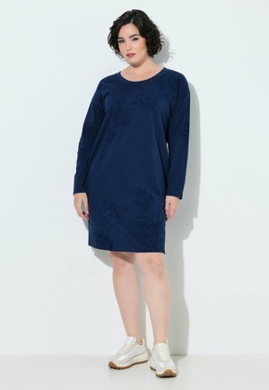 Vrouw met kort krullend donker haar, die een marineblauwe jurk met lange mouwen en witte sneakers draagt, staand tegen een lichte muur.