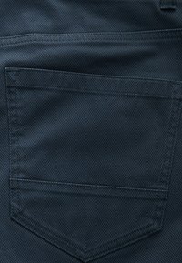 Dunkelblauer Stoff mit einem strukturierten Muster, der eine Gesäßtasche mit geformtem Saum und sauberer Naht aufweist. Keine sichtbaren Beschläge oder Akzente.
