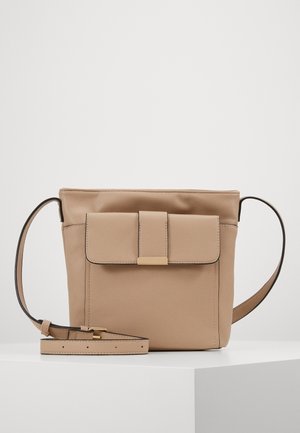 Beżowa skórzana torba na ramię z przednią kieszenią na klapę i prostokątnym zapięciem w kolorze złotym, regulowany pasek zwinięty na białej powierzchni.