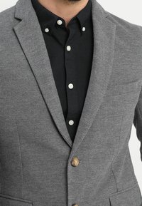 Szary żakiet teksturowany z klapami, posiadający zapięcie na jeden mosiężny guzik oraz czarną koszulę z guzikami pod spodem.