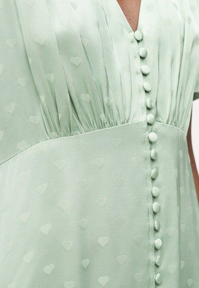 Κοντινό πλάνο σε ανοιχτοπράσινο φόρεμα με κουμπιά καλυμμένα με ύφασμα και διακριτικό ανάγλυφο σχέδιο καρδιάς σε ρυθμισμένο μπούστο και μέση.