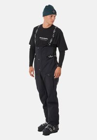 Schwarze Skihosen mit verstellbaren Trägern, ausgestattet mit Reißverschlusstaschen und einer taillierten Passform. Kombiniert mit einem schwarzen grafischen T-Shirt und einer blauen Mütze.