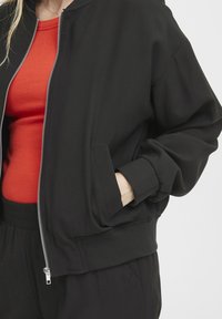 Čierna bomber bunda so zipsom, hrdlovými manžetami a bočnou kapsou. Skombinovaná s červeným pruhovaným vrchom pod ňou.