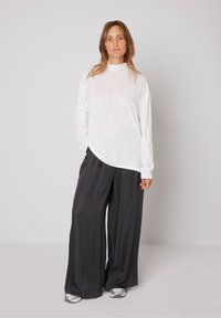Bijela bluza dugih rukava s visokim ovratnikom, u kombinaciji s labavim crnim hlačama, obje izrađene od mekog, laganog materijala. Model stoji protiv neutralne pozadine.