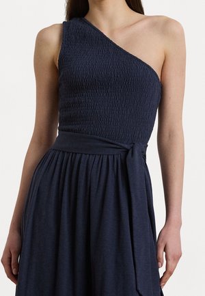 Femme portant une robe bleu marine asymétrique avec un haut texturé et une ceinture nouée à la taille, debout devant un fond uni.