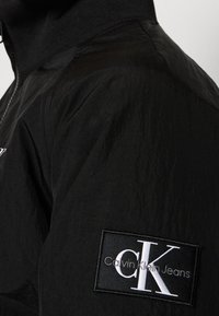 Veste noire zippée en tissu texturé. Elle présente un grand patch logo "Calvin Klein Jeans" sur le côté gauche. Col lisse.