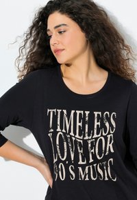 Černé, volné tričko s kulatým výstřihem, na kterém se nachází výrazný béžový grafický potisk s nápisem "NADČASOVÁ LÁSKA K HUDBĚ 90. LET".