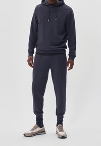 Felpa navy con cappuccio e coulisse, abbinata a pantaloni jogger a gamba affusolata coordinati. Le calzature presentano una combinazione di colori beige, grigio e bianco.