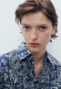 Camicia blu con motivo paisley, dotata di colletto e realizzata in tessuto testurizzato. Presenta disegni floreali bianchi e una vestibilità comoda.