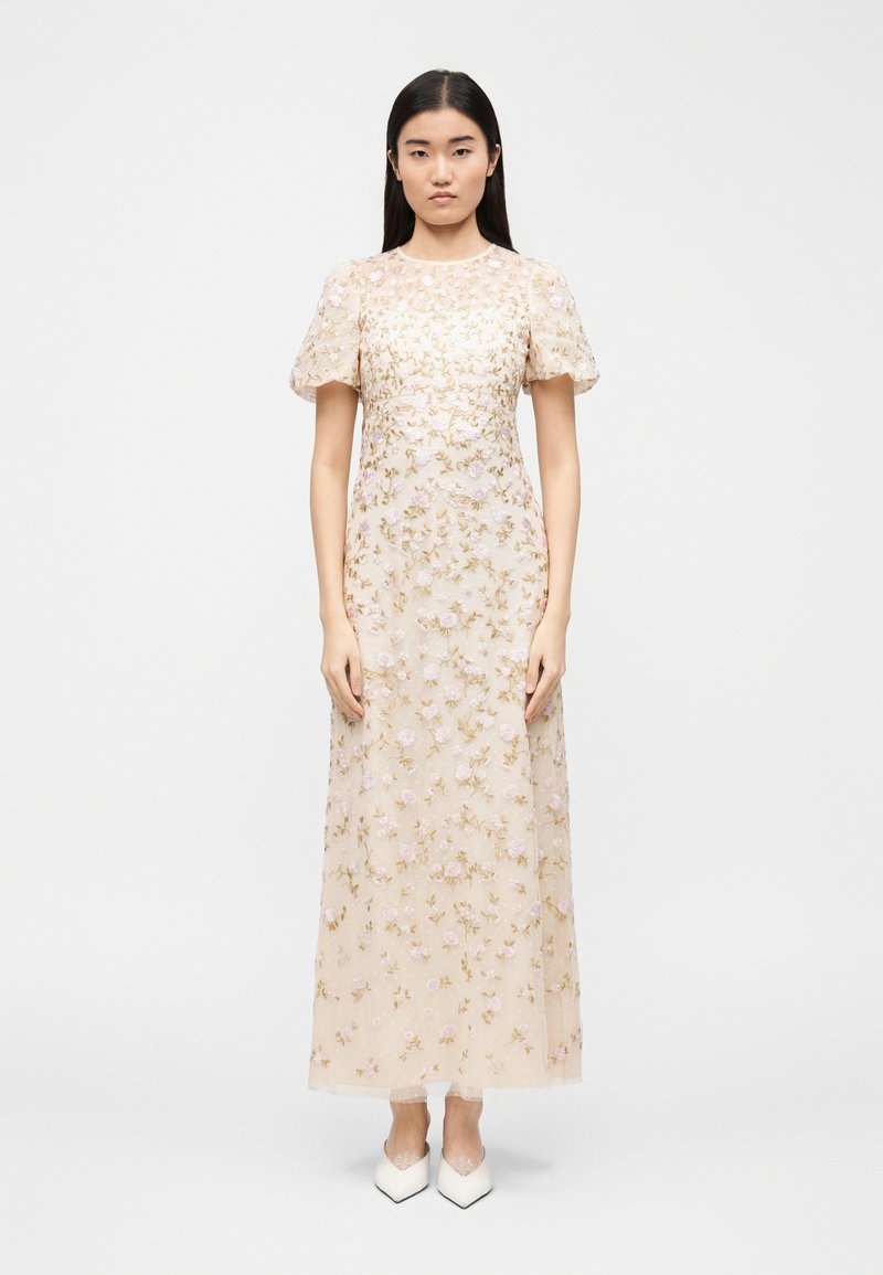 Γυναίκα με μακριά σκούρα μαλλιά, που φοράει ένα διάφανο, κρεμ χρώματος φόρεμα μέχρι το δάπεδο, κεντημένο με ρόζ και χρυσά ανθικά σχέδια, και λευκά μυτερά παπούτσια.