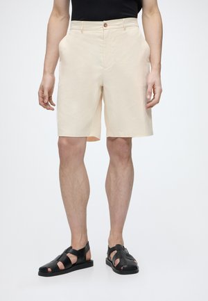 Homme portant un short beige jusqu'aux genoux, un haut noir sans manches et des sandales noires à bout ouvert, debout sur un fond blanc.