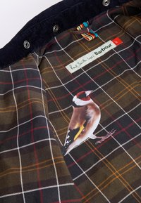 Εσωτερικό σακακιού από καστόρι με σκουρόχρωμο καρό σχέδιο σε αποχρώσεις καφέ, κίτρινου και λευκού, καθώς και μια απεικόνιση πτηνού στο ύφασμα.