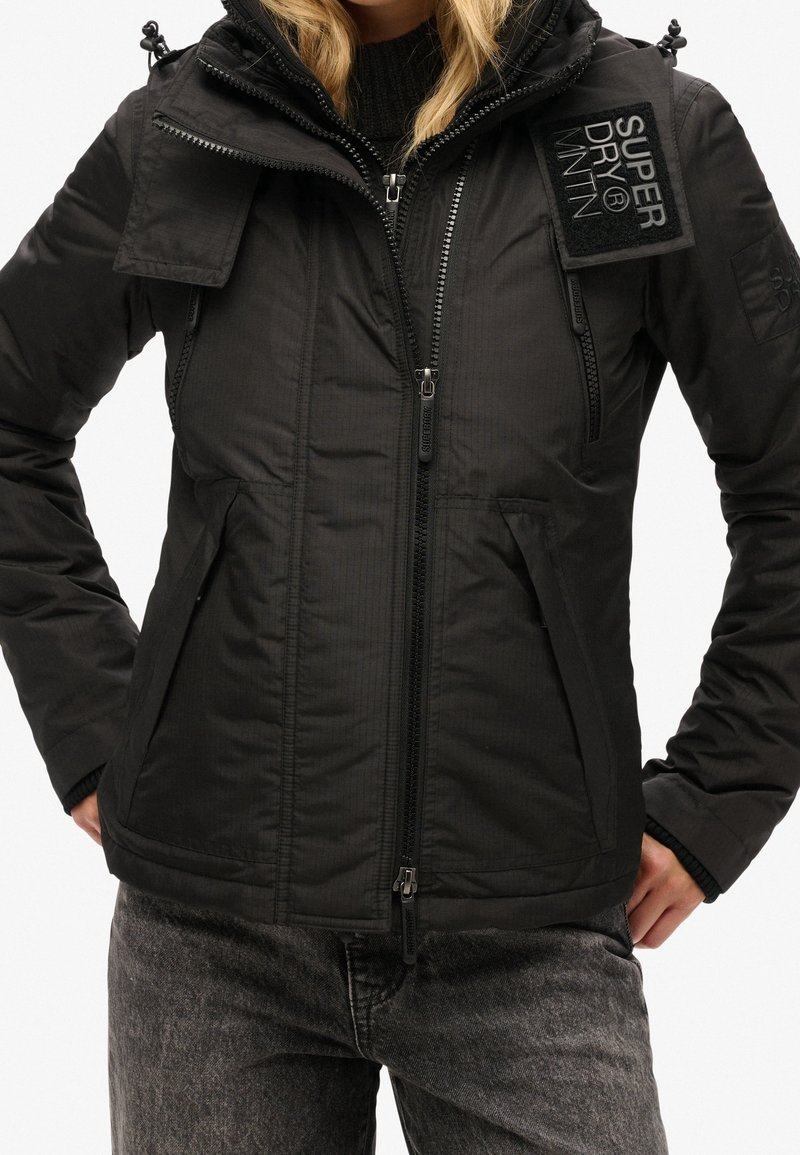 Schwarze isolierte Jacke mit durchgehendem Reißverschluss, mehreren Taschen und abnehmbarer Kapuze. Ausgestattet mit strukturiertem Stoff und Logopatch an der Schulter.