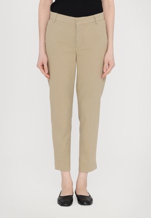 Mujer con pantalones beige de pierna recta y zapatos planos negros sobre un fondo blanco, con las manos descansando a los costados.