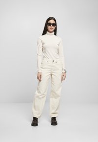 Top in beige chiaro a collo alto, jeans larghi bianco sporco e stivali stringati marrone scuro. Il modello indossa occhiali da sole neri; design e texture minimalisti.