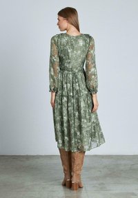 Vestido floral verde con mangas largas transparentes, cintura fruncida y falda en niveles. Combinado con botas beige hasta la rodilla. Tejido ligero y suave.