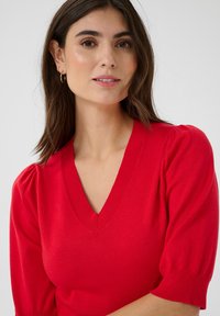 Punainen neulepaita, jossa on V-pääntie ja lyhyet puff-hihat. Suutarin sileä pinta ja istuva muotoilu korostavat elävää väriä ja hienostunutta yksityiskohtaa.