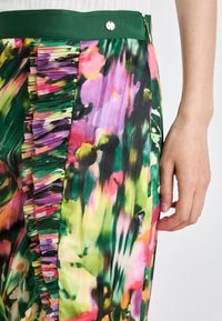 Kalhoty s květinovým vzorem mají plisé na bocích, zelený pas a živé barvy včetně růžové, fialové, zelené a žluté.