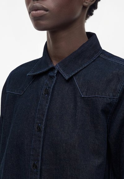 Chemise en jean foncé à boutons avec col pointu portée par une personne à la peau foncée et aux cheveux courts et bouclés, montrée du cou à la poitrine.