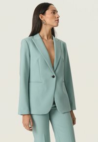 Világoskék, szabott zakó mély V-nyakkivágással, hosszú ujjakkal, egy gombos zárással és két elülső zsebbel. Sima, texturált anyag.