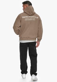 Brown hoodie with a large back print, featuring white text. Paired with black pants and white sneakers. Comfortable, loose fit.