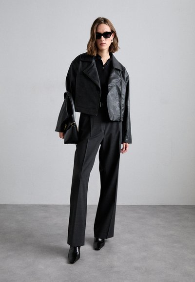 Giacca cropped in pelle nera sopra un top in maglia nera, abbinata a pantaloni grigi a vita alta e stivali neri a punta. Tiene una piccola borsa nera.