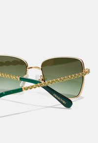 Zaļgandijas kvadrāta saulesbrilles ar zelta rāmi un zaļiem kristāla akcentiem uz kājiņām. Tām ir spīdīga zaļā kājiņa ar "Swarovski" zīmolu.