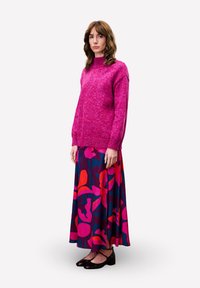 Femme avec des cheveux bruns ondulés portant un pull roses vif, une longue jupe fleurie colorée et des chaussures Mary Jane noires, se tenant devant un fond blanc.