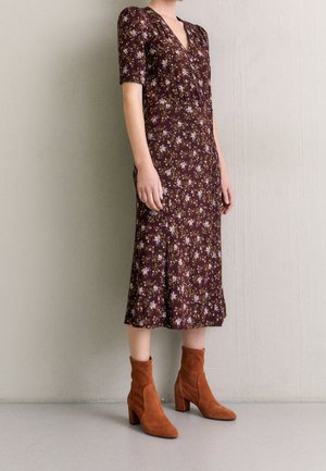 Osoba ubrana w brązową, kwiecistą sukienkę midi z krótkimi rękawami, w połączeniu z rdzawymi zamszowymi botkami za kostkę, stoi na tle jednolitej ściany.