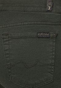Denim verde oscuro con una costura decorativa en el bolsillo trasero y una etiqueta negra que dice "para toda la humanidad". Presenta un botón metálico y líneas limpias.