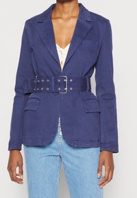 Blazer in cotone blu navy con una silhouette aderente, tasche frontali e una cintura larga abbinata con accessori in argento. Finitura in tessuto testurizzato.