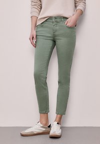 Donna che indossa pantaloni aderenti verde chiaro, maglione beige e scarpe da ginnastica bianche, in piedi contro una parete chiara con la mano in tasca.