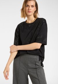 Czarny t-shirt z krótkim rękawem, ozdobiony cekinami na ramionach i górnej części przodu, w połączeniu z szarymi, luźnymi spodniami. Prosty, casualowy design.