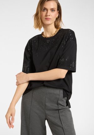 Schwarzes T-Shirt mit kurzen Ärmeln und Paillettenakzenten auf den Schultern und der oberen Vorderseite, kombiniert mit grauen, locker sitzenden Hosen. Einfaches, lässiges Design.