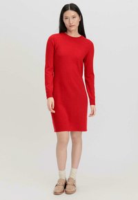 Abito a maglia lungo rosso con maniche lunghe e scollatura a girocollo, caratterizzato da una silhouette dritta e una texture morbida. Indossato con scarpe mocassino beige e calzini leggeri.