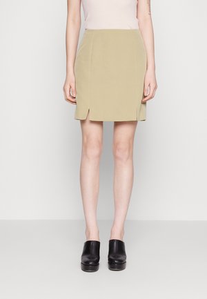 Ανοιχτό ελιά πράσινη μίνι φούστα με πλαϊνές σχισμές, ίσια κοπή, λεία ύφασμα, σε συνδυασμό με μαύρες ψηλές μπλοκ γόβες.
