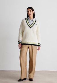 Weißer Rippstrickpullover mit V-Ausschnitt, marine- und gelbgestreiften Akzenten, kombiniert mit beigen Hosen und braunen Schuhen. Glatte Textur, lässige Passform.