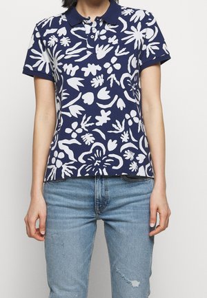 Kobieta ubrana w granatową koszulkę polo z białymi abstrakcyjnymi wzorami kwiatowymi oraz jasnoniebieskie, postrzępione jeansy, stojąca na jednolitym tle.