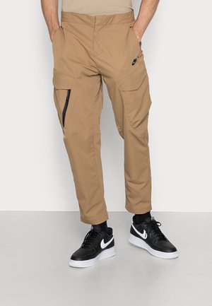 Mężczyzna w beżowych spodniach typu cargo z kieszenią na zamek i czarnych butach sportowych Nike z białą podeszwą.