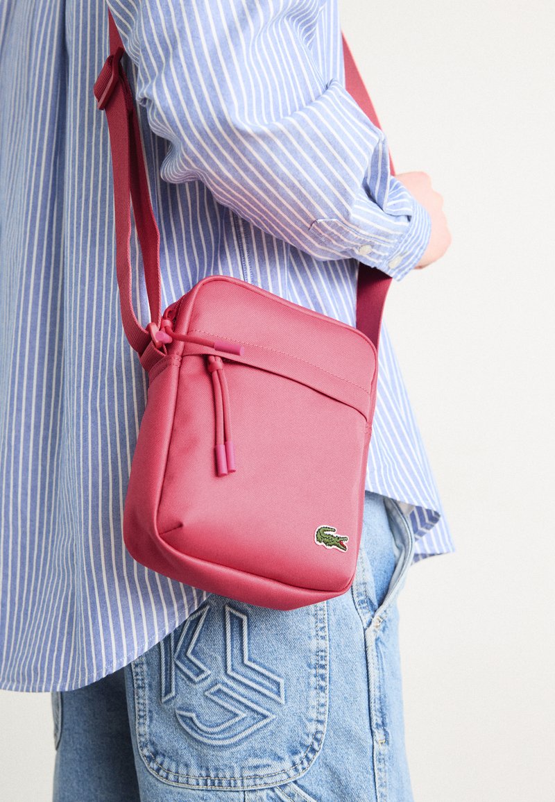 Sac en bandoulière en tissu rose avec un design plat, fermeture éclair sur le dessus, bandoulière ajustable et détail de logo brodé, porté sur une chemise rayée bleue.